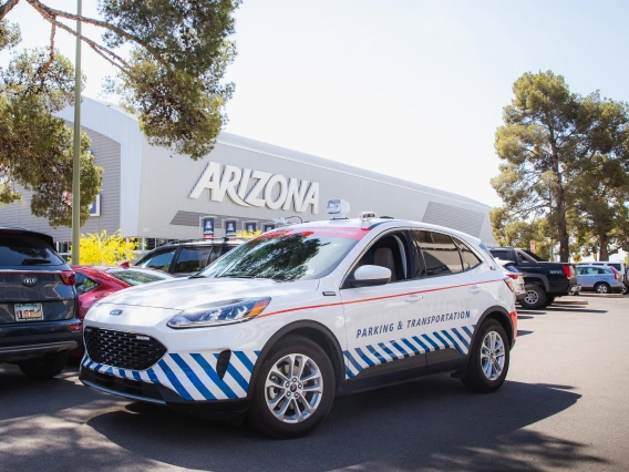 A University of Arizona Parking & Transportation vehicle, a white Ford SUV with blue diagonal stripes and red accents, is parked in a lot near the Arizona Stadium. The words "Parking & Transportation" are displayed on the side of the vehicle. The background features a large building with "ARIZONA" in bold letters, along with parked cars and tall trees providing shade. The scene is set on a bright, sunny day, highlighting the university's transportation services in action.