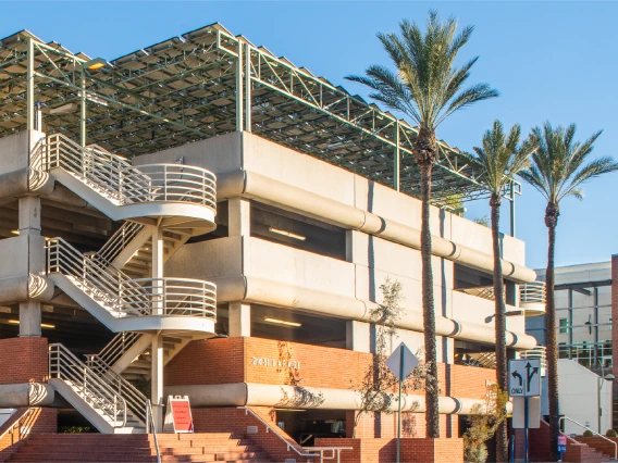 The Second Street Garage at the University of Arizona, a multi-level concrete parking structure with an external staircase, is shown on a sunny day. The garage features a solar panel canopy on the roof and is surrounded by tall palm trees. A traffic light and street signs mark the intersection of Mountain Avenue and Second Street in the foreground, with tram tracks running along the street. The Student Union Memorial Center, displaying the University of Arizona "A" logo, is visible to the right of the garag