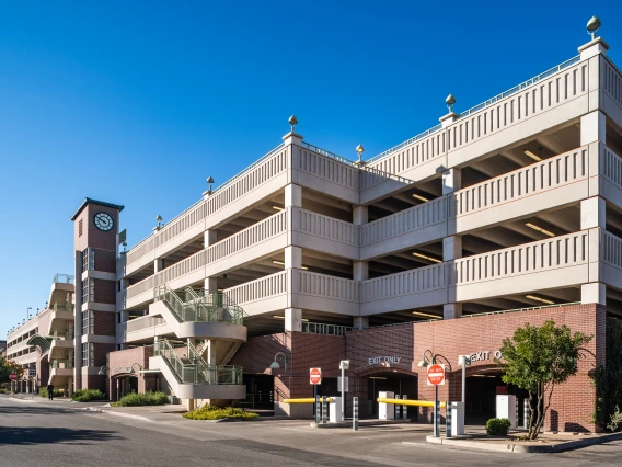 A multi-level Tyndall parking garage with a brick and concrete facade, featuring a clock tower on the left side. The structure has external staircases with green railings and multiple 'Do Not Enter' signs at the exit points. The sky is clear and blue, and the surrounding area includes some greenery and a few pedestrians.