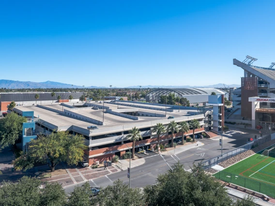 An aerial view of a multi-level cherry garage near Arizona Stadium at the University of Arizona. The structure has a brick and concrete facade with external staircases and palm trees lining the surrounding sidewalks. The stadium, with its large seating area and signage, is visible to the right, while a sports field with artificial turf is in the foreground. In the background, additional university buildings, a large arched structure, and a mountain range are visible under a clear blue sky.