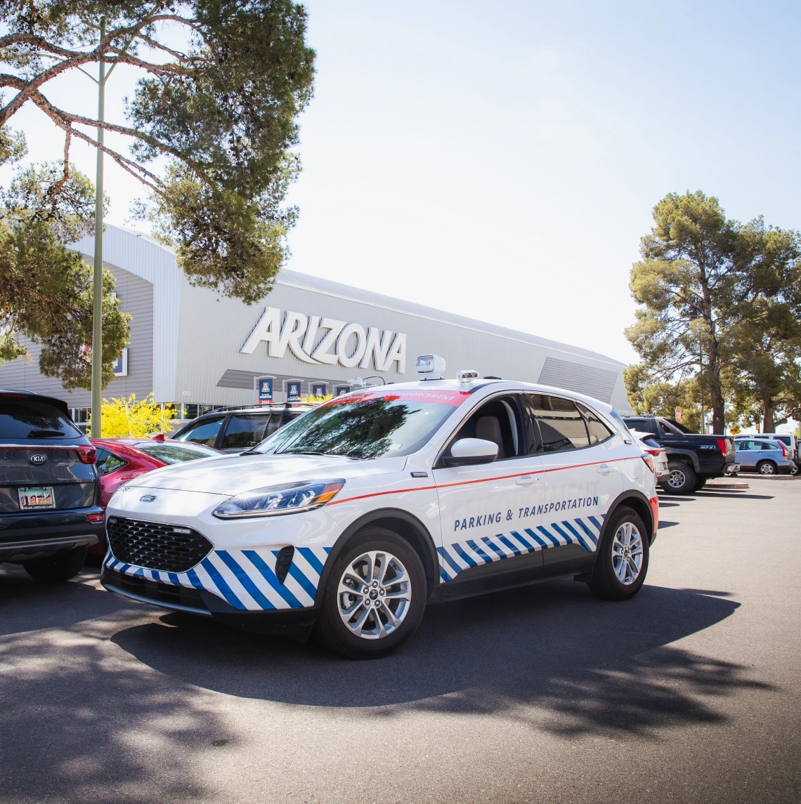 A University of Arizona Parking & Transportation vehicle, a white Ford SUV with blue diagonal stripes and red accents, is parked in a lot near the Arizona Stadium. The words "Parking & Transportation" are displayed on the side of the vehicle. The background features a large building with "ARIZONA" in bold letters, along with parked cars and tall trees providing shade. The scene is set on a bright, sunny day, highlighting the university's transportation services in action.