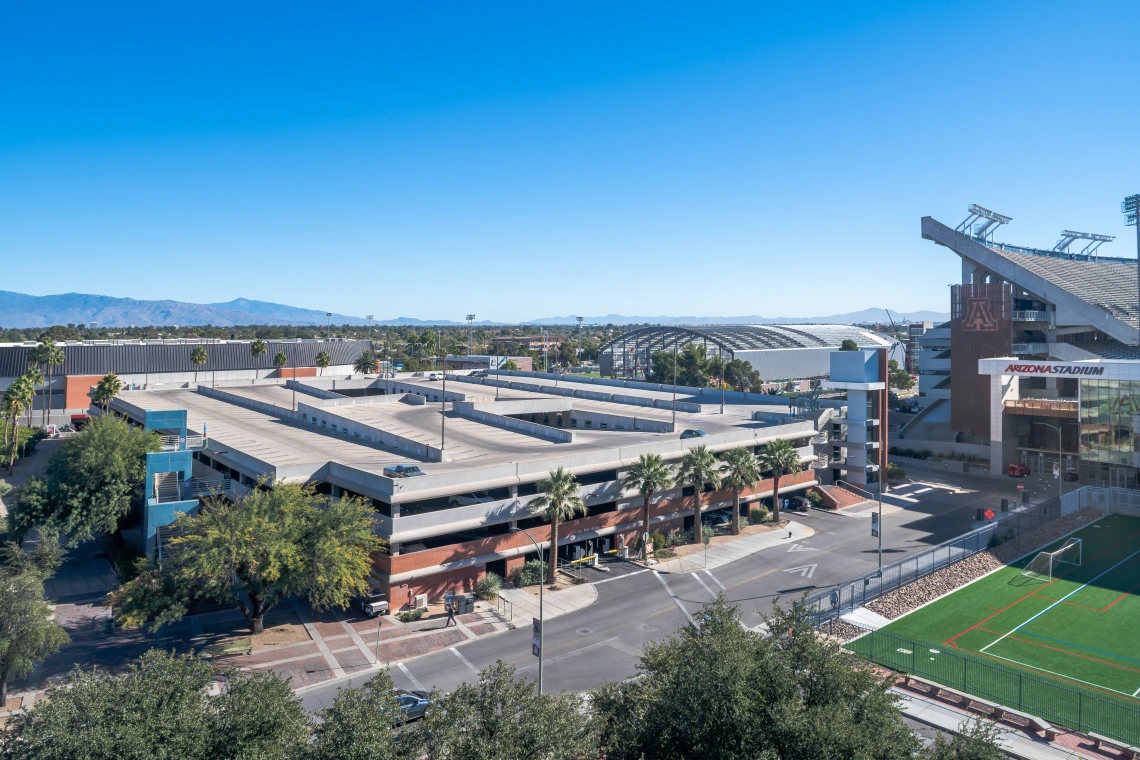 An aerial view of a multi-level cherry garage near Arizona Stadium at the University of Arizona. The structure has a brick and concrete facade with external staircases and palm trees lining the surrounding sidewalks. The stadium, with its large seating area and signage, is visible to the right, while a sports field with artificial turf is in the foreground. In the background, additional university buildings, a large arched structure, and a mountain range are visible under a clear blue sky.