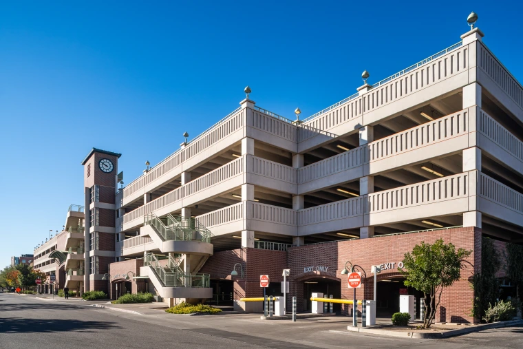 A multi-level Tyndall parking garage with a brick and concrete facade, featuring a clock tower on the left side. The structure has external staircases with green railings and multiple 'Do Not Enter' signs at the exit points. The sky is clear and blue, and the surrounding area includes some greenery and a few pedestrians.