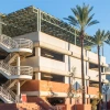 The Second Street Garage at the University of Arizona, a multi-level concrete parking structure with an external staircase, is shown on a sunny day. The garage features a solar panel canopy on the roof and is surrounded by tall palm trees. A traffic light and street signs mark the intersection of Mountain Avenue and Second Street in the foreground, with tram tracks running along the street. The Student Union Memorial Center, displaying the University of Arizona "A" logo, is visible to the right of the garag