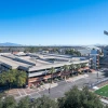 An aerial view of a multi-level cherry garage near Arizona Stadium at the University of Arizona. The structure has a brick and concrete facade with external staircases and palm trees lining the surrounding sidewalks. The stadium, with its large seating area and signage, is visible to the right, while a sports field with artificial turf is in the foreground. In the background, additional university buildings, a large arched structure, and a mountain range are visible under a clear blue sky.